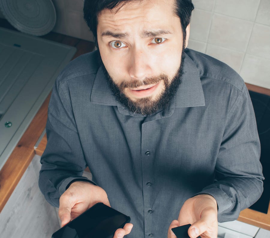 A confused man indoors looks up while holding a smartphone and credit card, concerned about a financial issue.